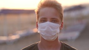 Closeup view video portrait of young white kid wearing medical face mask while standing outdoor in summer beach. Boy looks into camera.