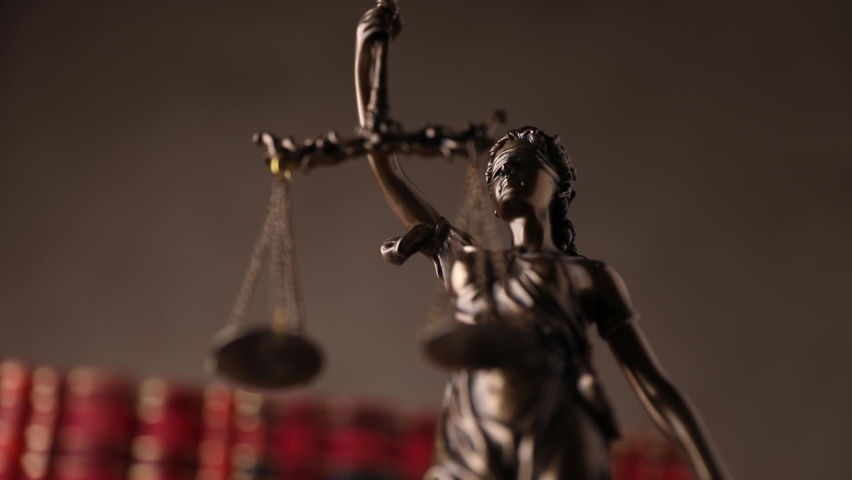 bronze law statue represented by blindfolded goddess spinning and holding a balance signifying impartiality and two broad sources of law in front of books on brown background Royalty-Free Stock Footage #1060234070
