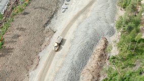 Aerial video on one heavy industrial yellow dump truck drives load of gravel on new highway road construction site, sunny day, pine tree forest around. 