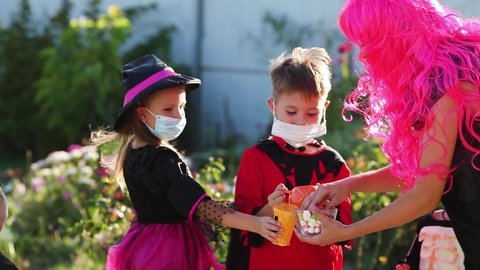 Children trick or treat in Halloween costume and medical mask. A little boy, girl and baby in suits during the coronavirus pandemic receive candy from a woman in a pink wig.