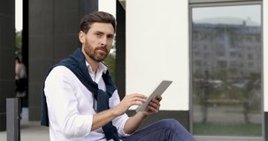 Attractive man with beard greeting someone with hand while sitting outdoors with digital tablet. Concept of people, gesturing and modern technology.