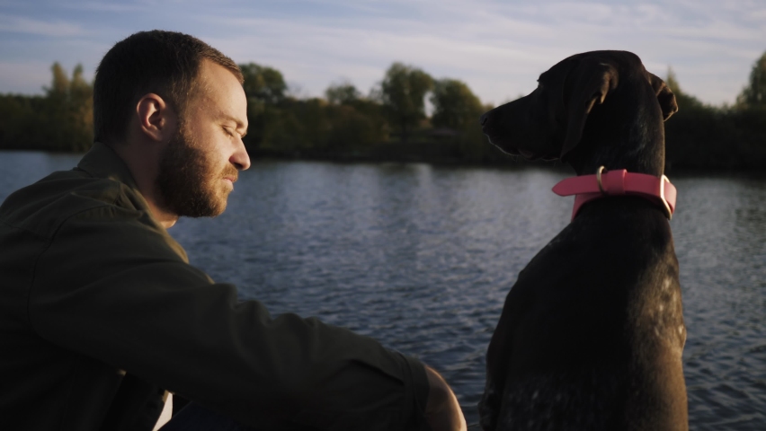 Side view of a young guy sitting with his pet dog on the pier and admiring the beauty of the autumn lake. A man looks kindly into the eyes of his dog.