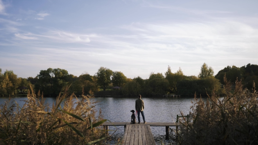 An amazing shot of a young man with his pet dog standing on the edge of the pier and admiring the beauty of the lake at sunset.