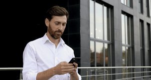 Cheerful bearded businessman in white shirt answering on emails with help of smartphone while walking on street. Concept of masculinity, confidence and technology.