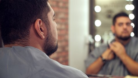 Bearded man himself grooming beard with trimmer front mirror in barbershop. Arabian man using electric razor for shaving beard in male salon
