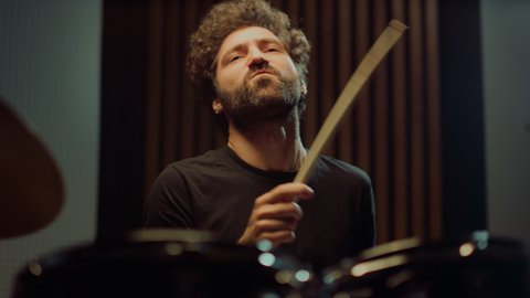Expressive artist nodding head in concert hall. Emotional drummer hitting drum cymbals in repetition studio. Curly male musician making rock music indoor.