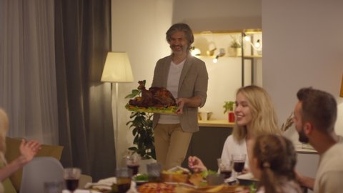 Happy middle-aged man bringing big roasted turkey to Thanksgiving day dinner, his cheerful family clapping hands, slowmo shot footage