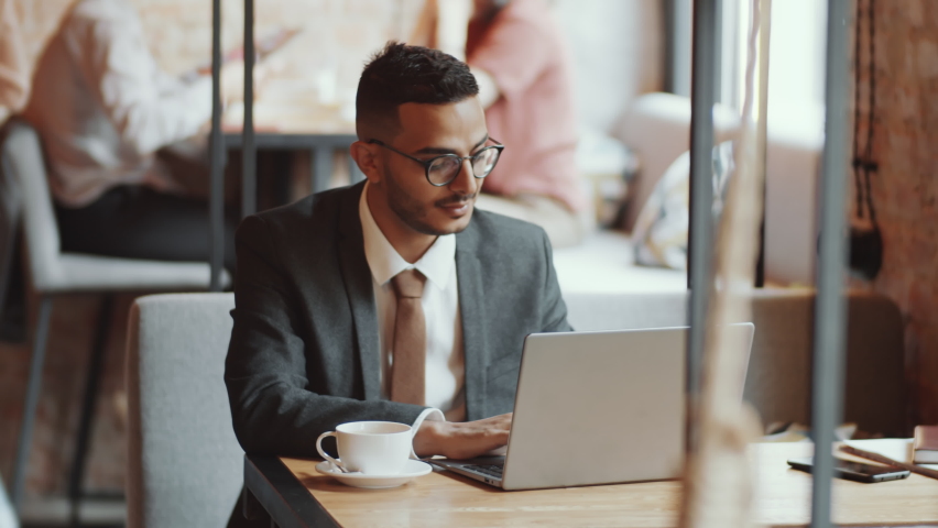 Handsome middle eastern businessman in formalwear and glasses typing on laptop and then looking at side thoughtfully while working at cafe table Royalty-Free Stock Footage #1060256786