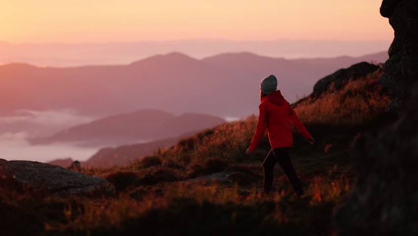 Young woman in orange jacket running up on top of mountain summit at sunset, raises arms into air, happy and drunk on life, youth and happiness. Watching the sunset with beautiful landscape | Shutterstock HD Video #1060258418