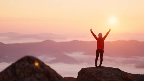 Young woman in orange jacket running up on top of mountain summit at sunset, raises arms into air, happy and drunk on life, youth and happiness. Watching the sunset with beautiful landscape