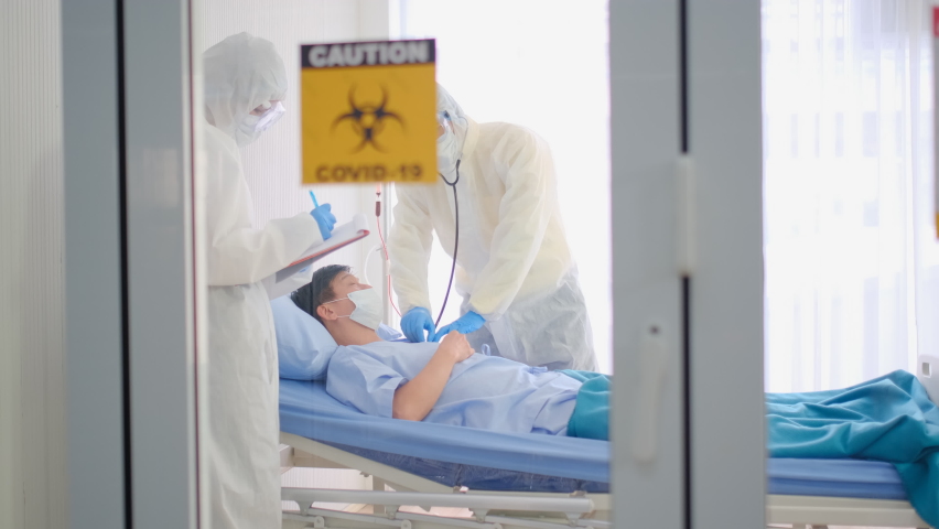 Doctor with full covered body use stethoscope to analysis sign and symptom of Covid-19 infected patient in closed room with biohazard banner. Nurse with PPE suit also record the data in the room.  Royalty-Free Stock Footage #1060259291