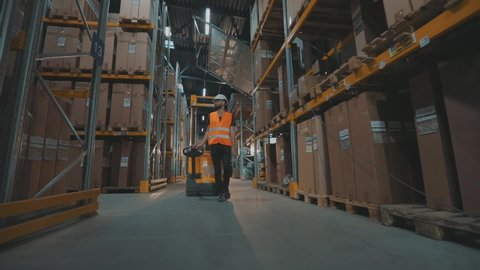 A worker transports cargo through the warehouse. A worker walks through the warehouse with an electric hydraulic forklift.