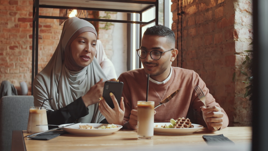 Young muslim woman in hijab sitting with boyfriend at cafe table, eating lunch and discussing something on smartphone | Shutterstock HD Video #1060269767
