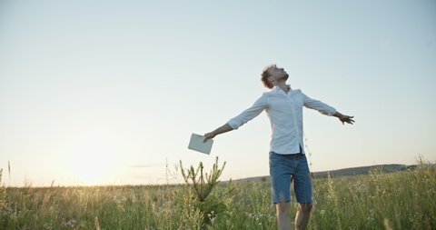 Low angle of young male poet making notes and thinking then spinning around with outstretched arms at sunset in meadow