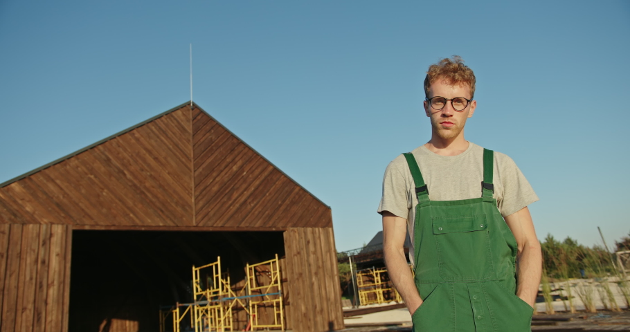 Low angle of confident man in uniform and glasses crossing arms and looking at camera while standing outside wooden shed in countryside