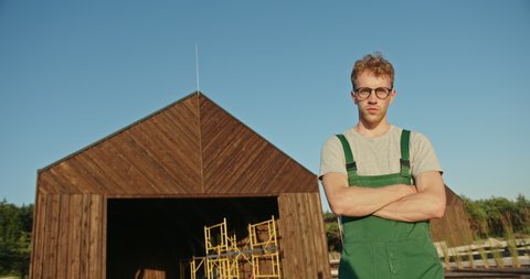 Low angle of confident man in uniform and glasses crossing arms and looking at camera while standing outside wooden shed in countryside