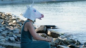 Upset woman with unicorn mask sitting by the sea, strange weird video.
