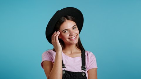 Attractive hipster playful woman in overall wear and hat smiling, flirting to camera over blue wall background. Cute trendy girl's portrait
