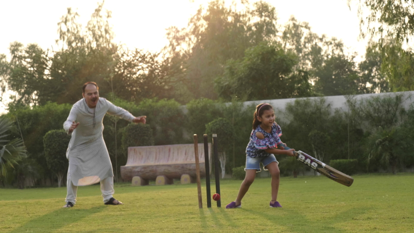 A young active girl child holding a bat and tries to hit a ball while playing cricket with the family members. A grandfather playing outdoor sport with granddaughter in a garden or recreational park Royalty-Free Stock Footage #1060275743