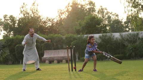 A young active girl child holding a bat and tries to hit a ball while playing cricket with the family members. A grandfather playing outdoor sport with granddaughter in a garden or recreational park