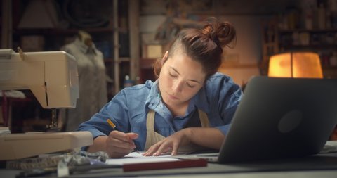 Portrait of confident modern young woman designer working on project in cozy workshop, beautiful pensive ginger female artisan drawing using pencil fashion design on paper work-desk indoors