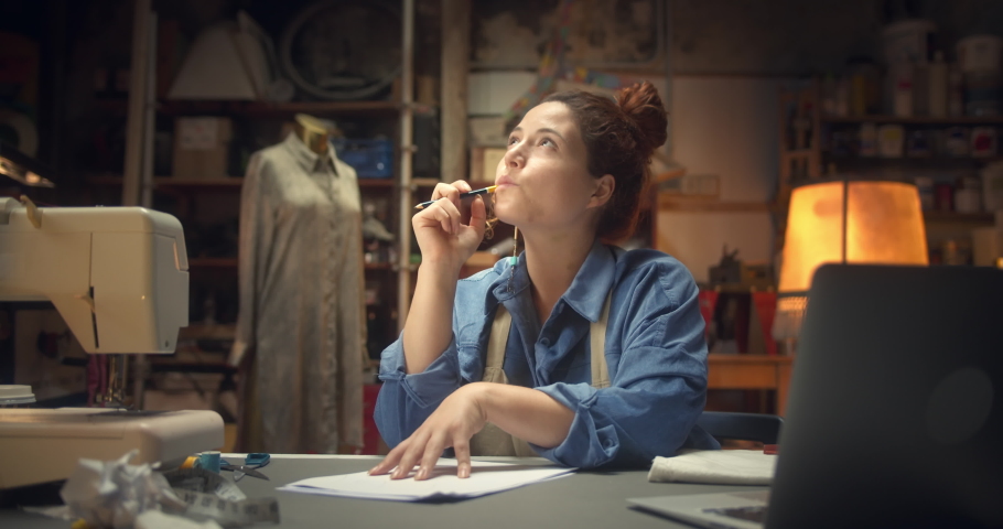 Beautiful young woman fashion designer getting inspiration genial idea in cozy workshop, pretty ginger female artisan working sitting next to work-desk holding pencil looking pensive indoors Royalty-Free Stock Footage #1060285001