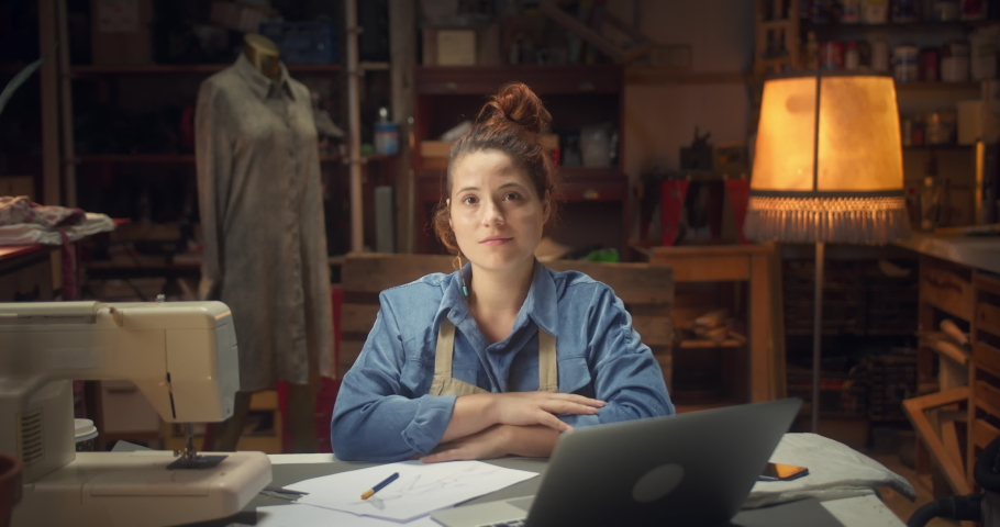 Portrait of cute young female designer sitting next to desk looking to camera in workshop, pensive pleasant artisan creative woman at workplace resting indoors with sewing machine and laptop on table