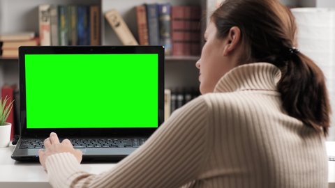 Woman sitting at table in apartment or office, she looks at green screen laptop and runs her index finger over laptop touchpad. Chroma key computer screen woman working on computer