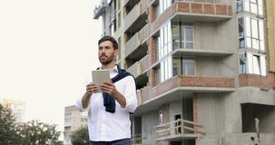 Attractive bearded man standing among unfinished construction of high-rise building and using digital gadget. Concept of people and technology.