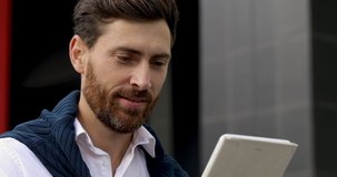 Close up of young man with stylish haircut and beard smiling while looking at digital tablet screen. Handsome guy in white shirt enjoying leisure time on fresh air.