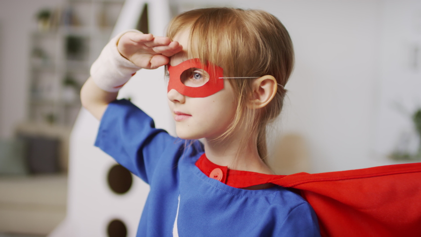 Medium closeup of small girl in superhero costume with red cape waving looking into the distance with cardboard rocket in background | Shutterstock HD Video #1060290539