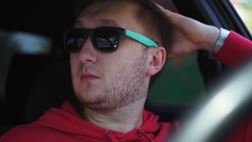 Guy sits car red T-shirt, hoodie, metal watch, sunglasses green souls. Put hand, looks out window, waits passenger, his wife, ponders, thinks, worries, chases thoughts, takes off glasses from his face.