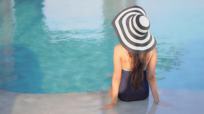 Back of Classy Female With Summer Hat in Swimsuit Sitting on Poolside of Luxury Pool. Static Full Frame