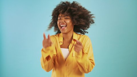 Young ethnic black female in casual outfit is shaking her hair, laughing and pointing at you by forefingers while posing against blue studio background. Close up, copy space, slow motion