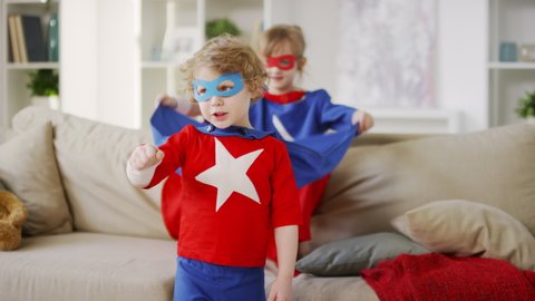 Medium shot of young boy in superhero costume with blue eye mask imitating flying like hero with fists forward and older sister sitting on sofa waving cloak in living room