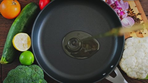 Cook pours sunflower oil into a frying pan, top view. Kitchen pan stands on brown boards, vegetables around it, and oil is poured into it for frying.  Slow motion  of  oil pouring into a frying pan. 