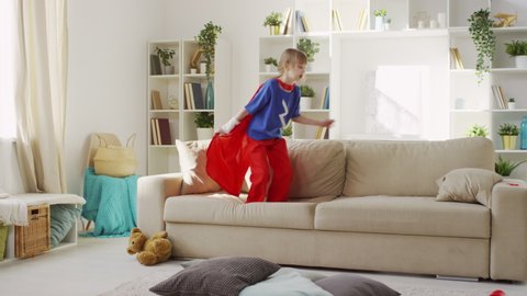 Slowmo follow shot of young girl in superhero costume jumping on sofa and waving cloak finally landing on floor with pillows laughing in living room