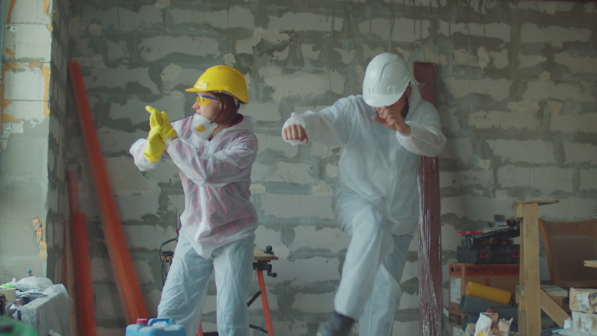 Young funny female and male worker in white protective suits dancing at construction site. Positive house renovation process. People in protective hardhats.  Royalty-Free Stock Footage #1060302368