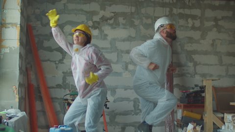 Young funny female and male worker in white protective suits dancing at construction site. Positive house renovation process. People in protective hardhats. 