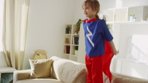 Slowmo follow medium shot of little girl in superhero costume jumping on sofa looking at camera and laughing in living room