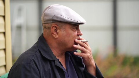 Close up of mature man smokes, sitting on bench in garden. Older male resting and smoking cigarette outdoors.