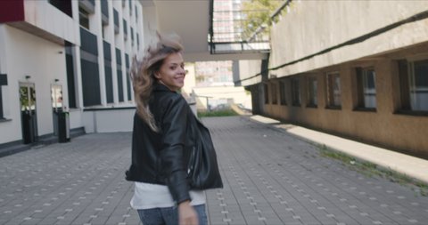 Young fashion woman flirting on background of urban street of city. Mixed race girl winks and invites to follow her. Video hand-held. Outdoors, lifestyle.