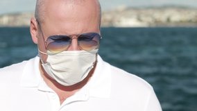 Video, an adult man in a white shirt and sunglasses removes and puts on a protective white mask. On a sunny day in summer, against the backdrop of the turquoise sea and sailing ships.