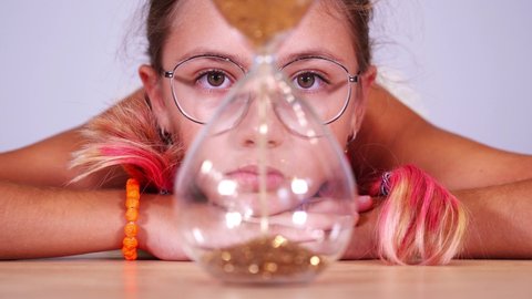 Girl with pink hair looks at sand inside sandglass at table