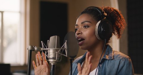 African american female musician in professional headphones singing song to mic at sound record studio, slow motion