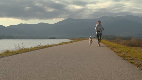Young woman running with dog on leash slow motion with mountains in background. Sports concept, animals concept.