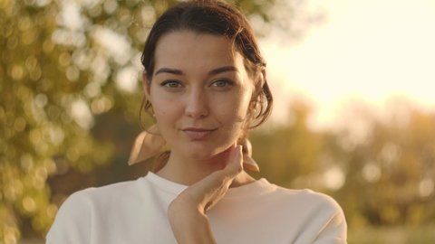 Portrait of a romantic brunette woman standing in the park at fall on a sunny day smiling and looking to the camera. The face of a happy beautiful woman in autumn outdoor at sunset. Slow-motion 4k
