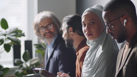 Tilt up shot of young muslim woman in hijab sitting with diverse male candidates in office, filling application form and then posing for camera while waiting for job interview