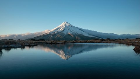 Time lapse: outdoor nature wilderness remote scene of white clouds billowing and flowing down Taranaki snow capped mountain peak by glass mirror reflective lake on blue sunny cloudless sky day