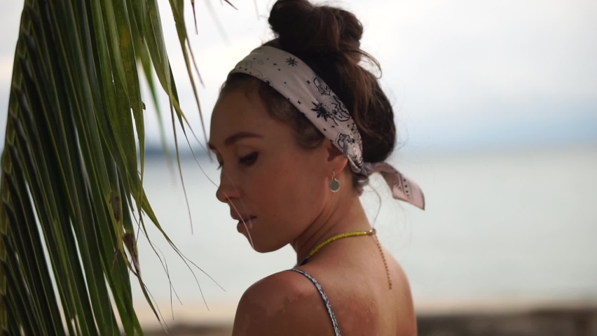 Beautiful young woman relaxing on a tropical beach under a palm tree at sunset.